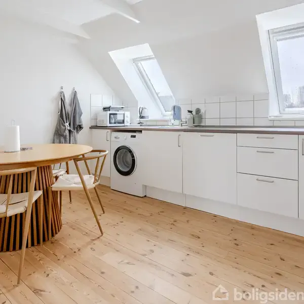 Rundt spisebord med stole stående på lyst trægulv. Moderne køkken med hvidt skab og vaskemaskine under skråvindue. Klar og lys atmosfære med enkelt design.