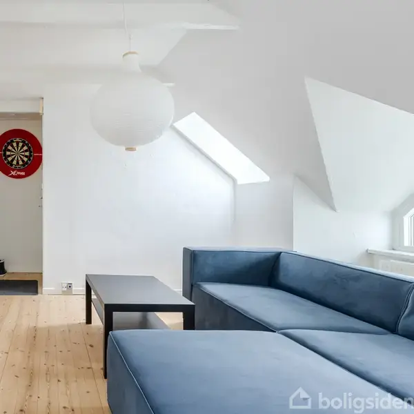 En blå sofa står i en stue under et skråt loft. Et bord står foran sofaen, og en dartskive hænger på væggen i baggrunden. Rummet er lyst med trægulv.
