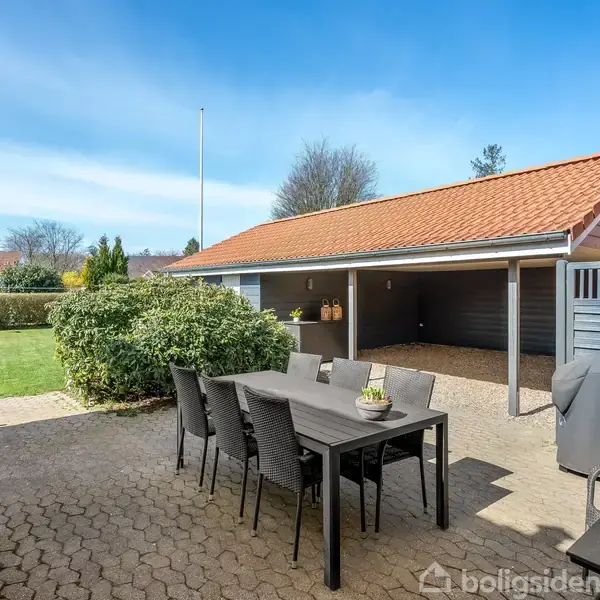 En sort spisebordssæt står stille på en flisebelagt terrasse. Omkranset af en velplejet græsplæne, tæt buskads og en dobbeltdækket carport med rødt tegltag i baggrunden.