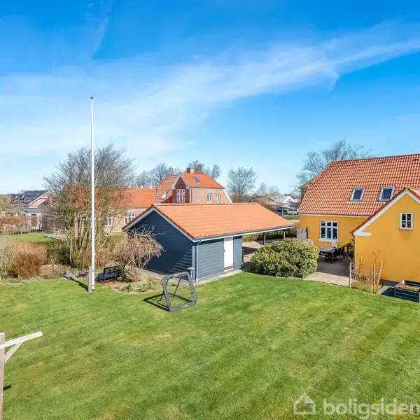 Gulmalet hus med rødt tag står ved siden af et gråt skur i en velholdt have. Græsplænen er omgivet af træer og buske, under en blå himmel uden skyer.