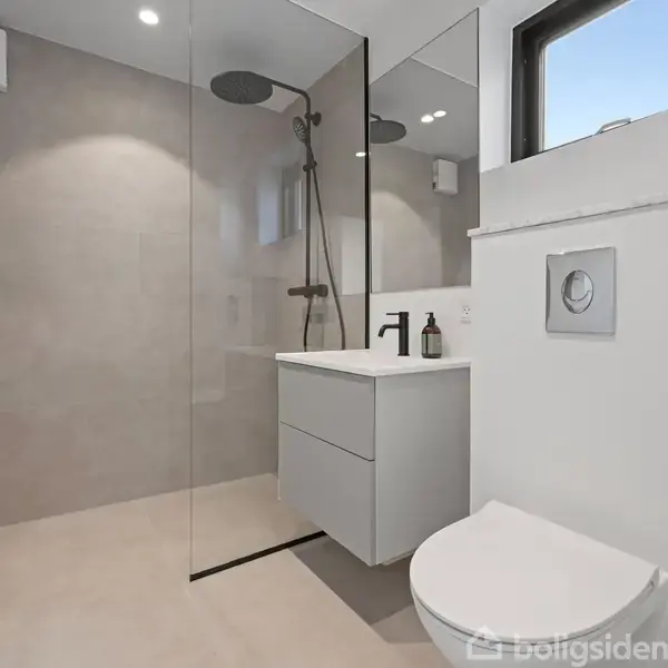 Bruser og vaskestativ står i et moderne badeværelse med glasvæg, grå fliser, væghængt toilet, og et lille vindue. En dispenser står ved vasken.