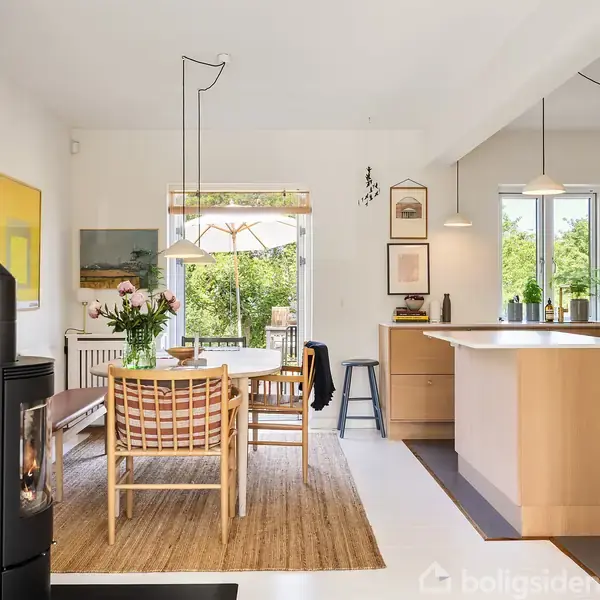 Stue med grå sofa, sofabord med gul vase, og lænestol ved siden, oplyst af bordlampe. Vægkunst inkluderer "LOUISIANA" og "POUL GERNES." Store vinduer udsigt til grøn natur.