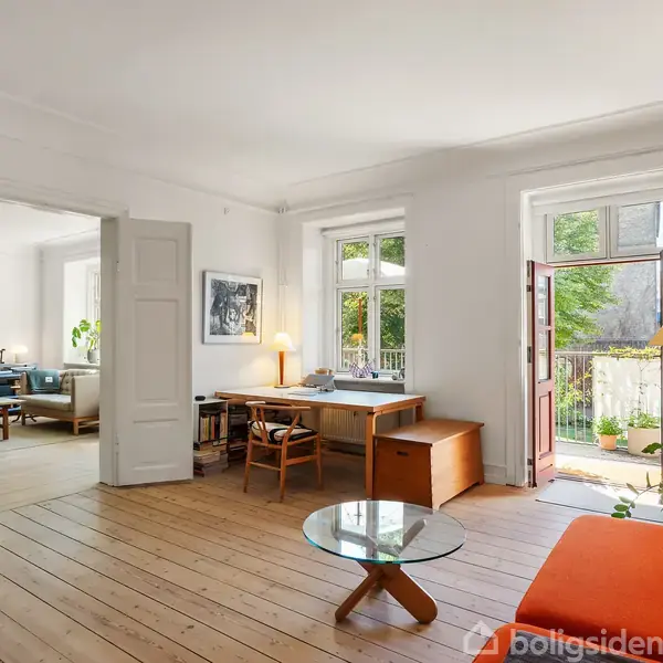 Stue med træplankegulv, hvor en orange sofa og glasbord står i forgrunden. Et skrivebord er placeret ved vinduet, der vender mod en lille altan med planter udenfor.