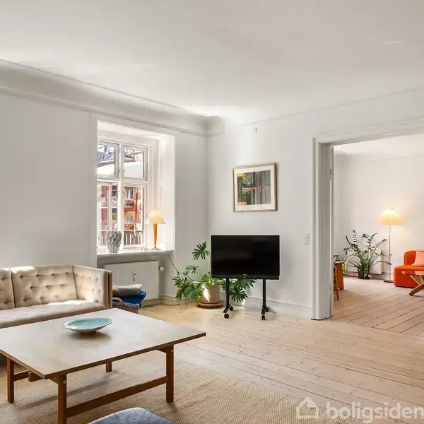 Stue med sofa, sofabord og fladskærms-TV. Rummet er lyst med trægulve, planter og store vinduer. I baggrunden ses en døråbning til et andet værelse med en orange stol og lampe.