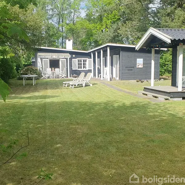 Et sort, træbeklædt sommerhus ligger i en grøn have med græsplæne. To liggestole står på plænen foran huset. Træer omkranser området.