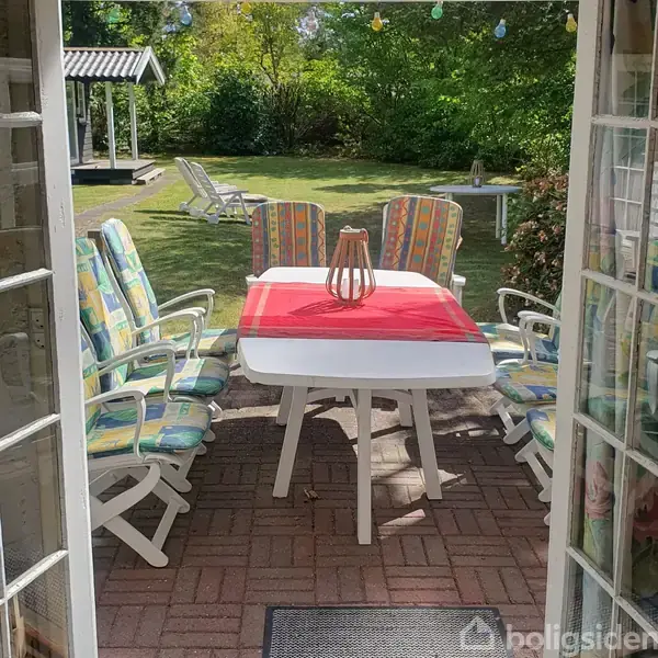 Et hvidt havebord med farverige stole er dækket med en rød dug og en trædekoration ovenpå. Det står på en terrasse med udsigt til en græsplæne og haveskur.