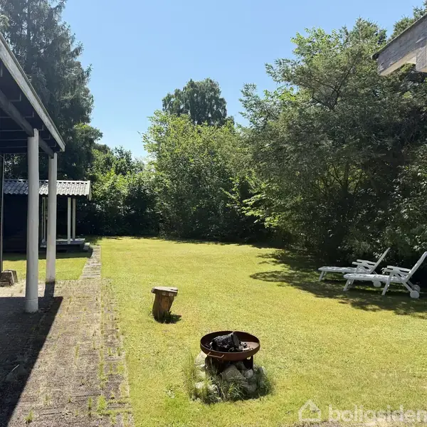 Stue med trævægge og -loft, brændeovn brænder ved siden af sofa og sofabord. Store vinduer til venstre med udsigt til træer. Malerier hænger på væggene.