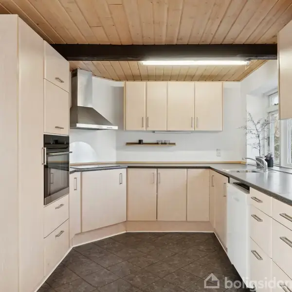 Et moderne køkken med lyse træskabe og mørke bordplader. Indbygget ovn og emhætte ses, mens store vinduer giver udsigt til naturen. Flisegulv og træloft fuldender rummet.