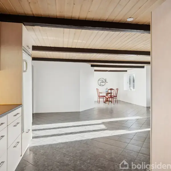 Et lyst køkken med hvide skabe og sorte bordplader fører til en spiseplads med et bord og fire stole. Loftet har synlige bjælker, og gulvet er flisebelagt.