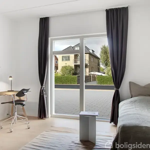 Et enkeltværelse med en seng til højre og et skrivebord med lampe og stol til venstre. Store vinduer med gardiner viser en udsigt til en boligbygning udenfor.