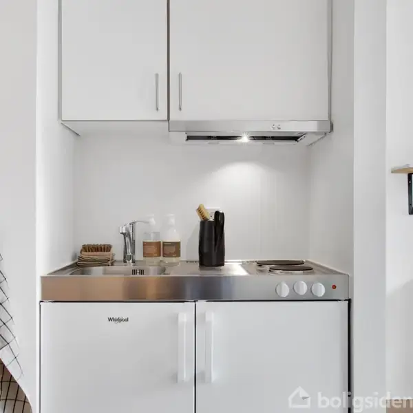 Et lille køkken med en Whirlpool ovn og kogeplade, hvide skabe, og en opvaskebørste ved siden af en krukke. En ternet klud hænger til venstre. Hylde til højre.