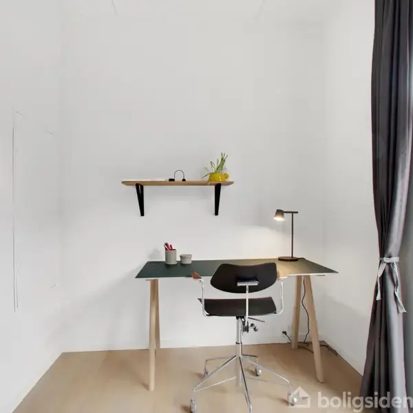 Et skrivebord med en kontorstol under en hylde hænger på en hvid væg. En lampe og små genstande står på bordet. Et vindue med gardiner ved siden af.
