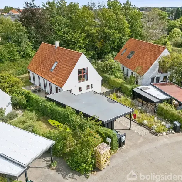 Hvide murstenshuse med røde tage står i en række i et grønt, beboelsesområde. En bil er parkeret i en indkørsel. Lyst grønt løv omkranser husene.