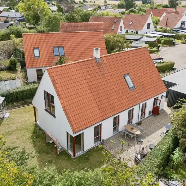 Huset har en rød tag og hvid facade. Det står i en have med græsplæne, flisebelagt terrasse og havepavillon. Flere lignende huse ses i det nærliggende villakvarter.
