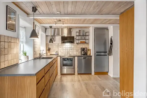 Et køkken med træskabe og grå bordplader, hvor opvaskemaskine, komfur og køleskab er integreret. Rummet er lyst med fliser på væggen og træ på gulv og loft.