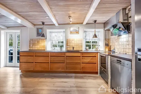 Køkken med træskabsfronter og stålapparater. To vinduer med gardiner, flisevæg, og to hængelamper. Trægulv og -loft. Dør til venstre fører udenfor. Lys og rummelig omgivelse.
