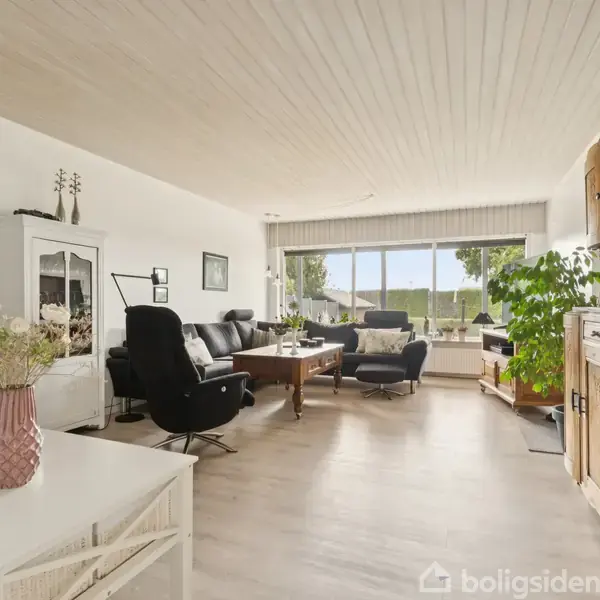 Stue med sorte lænestole og en sofa omkring et sofabord, lyse vægge og trægulve. Store vinduer til haven giver naturligt lys. Planter og træmøbler skaber en hyggelig atmosfære.
