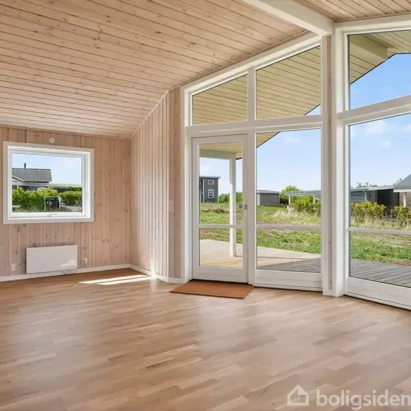 Stuen har træbeklædte vægge og lofter, store vinduer og en glasdør åbner ud til en træterrasse. Udenfor ses en grøn have og enkelte nabohuse.