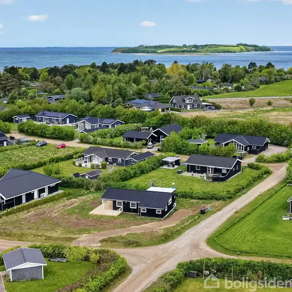 Flere sorte huse med græsplæner og grusveje omgiver et grønt landskab nær havet, med en ø i det fjerne. Klar blå himmel og spredte skyer ses over området.