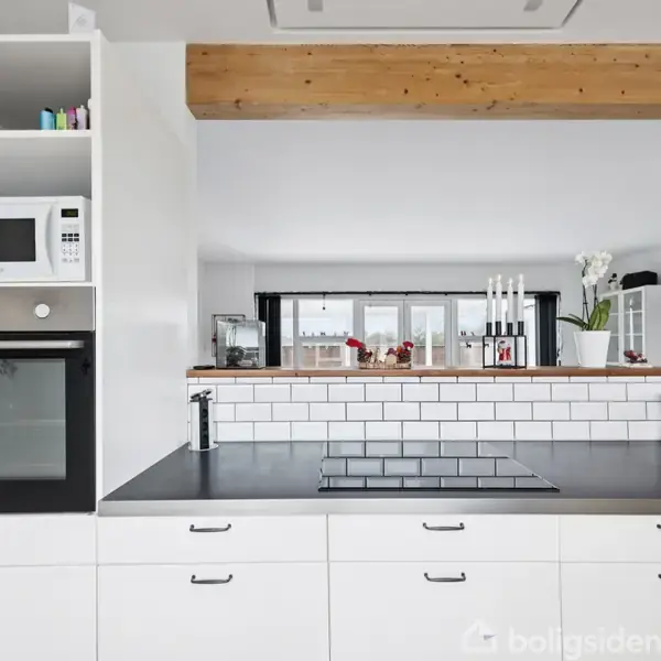 Køkkenbord med hvid bordplade og indbygget kogeplade, omgivet af hvide skabe. Til venstre er en mikroovn over en ovn. Bagved ses et vindue og dekorative genstande.