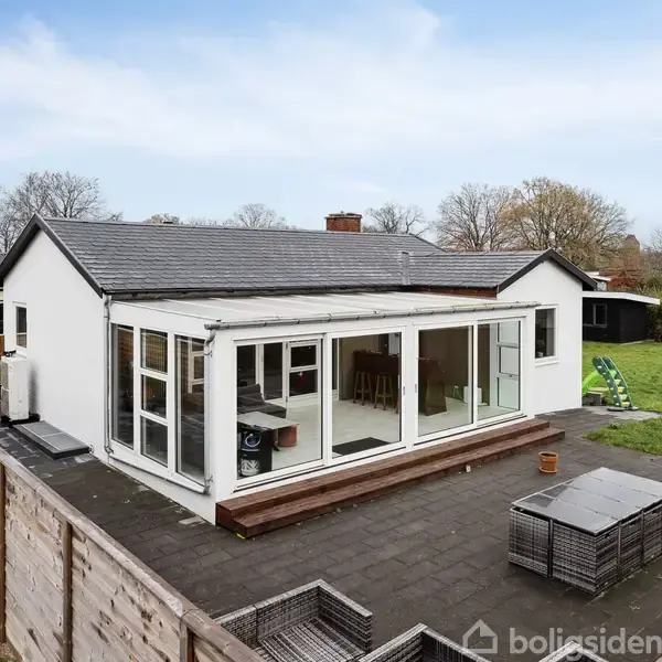 Et hvidt enfamiliehus med en stor glasinddækket veranda står i en have omgivet af højt hegn. Der er flisebelagt terrasse og græsplæne med legetøj i et roligt kvarter.