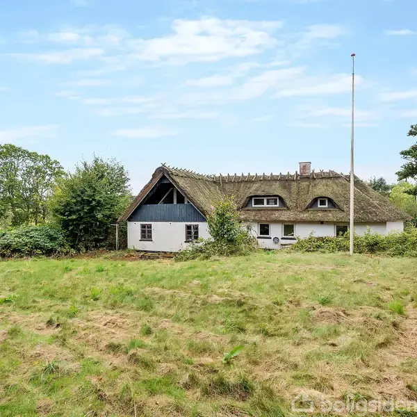 Et bindingsværkshus med stråtag står i en grøn have med en flagstang. Omkring huset er der tæt vegetation. I baggrunden ses åbne marker og en blå himmel med skyer.