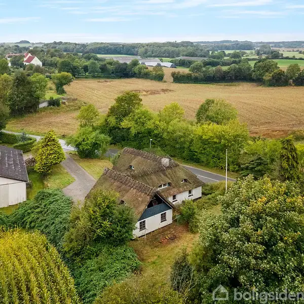 Hus med stråtag ligger i en landlig omgivelser omgivet af marker og træer. Der er en vej, der snor sig forbi, og en anden bygning ses tæt ved.