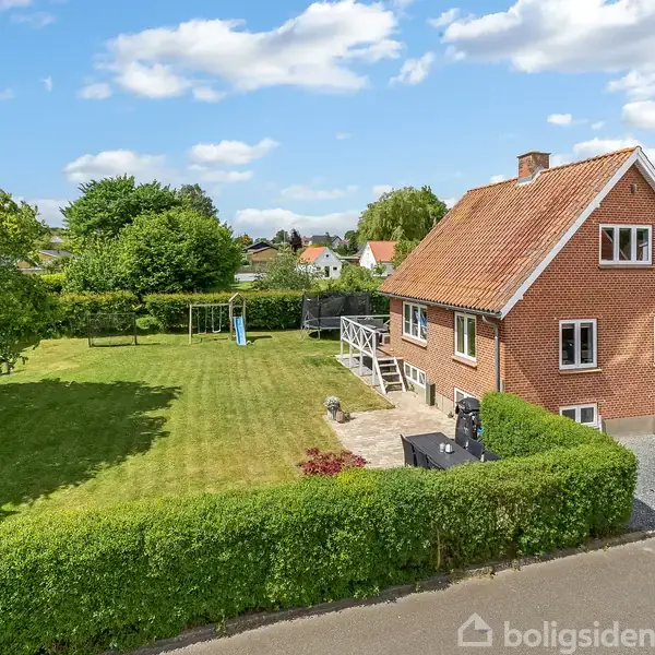En murstensvilla med rødt tegltag står midt i en velplejet have. En have med græsplæne omgivet af træer og hække, under en blå himmel med spredte skyer.