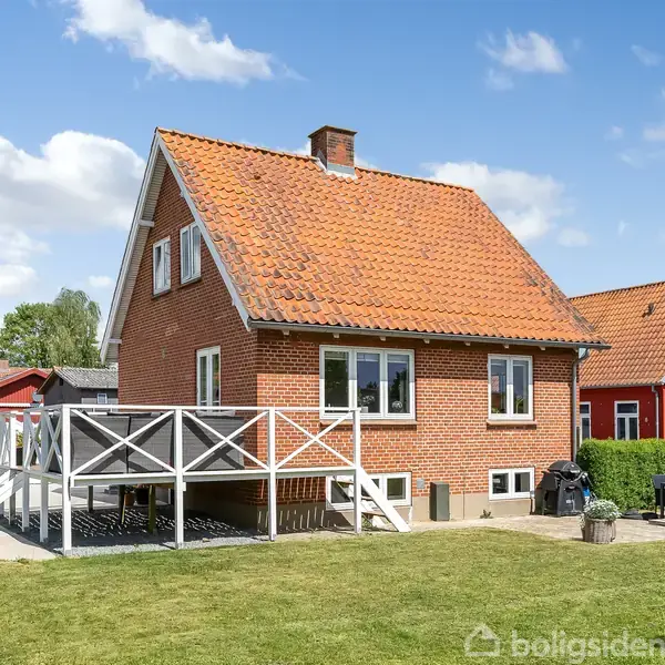 Rødt murstenshus med rød tagstenstag står i en velplejet have under en blå himmel. Til venstre er en terrasse med hvidt rækværk, og til højre ses havemøbler.