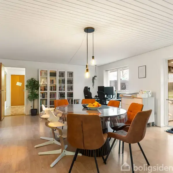 Rundt spisebord med stole omkring det, opstillet i en lys spisestue med trægulv. Bogreoler langs væggen, planter giver liv. Store vinduer slipper dagslys ind.