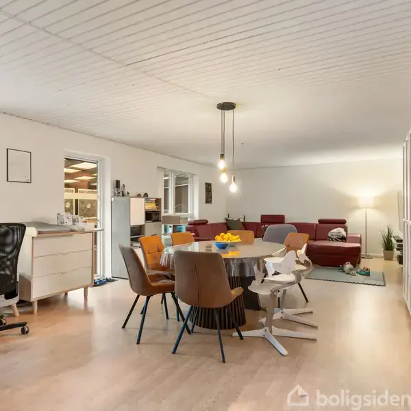Spisebord omgivet af stole står centralt. En sofa står i baggrunden. Skriveborde med computere ses til venstre. Rummet er lyst med trægulv og minimalistisk indretning.