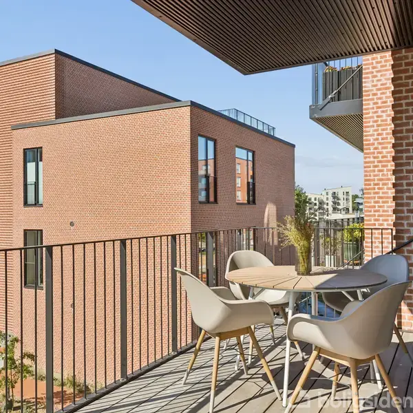 Et moderne altanbord med fire stole står stille på en altan. Omgivelserne er urbaniserede med murstensbygninger, og himlen er klar med få skyer.