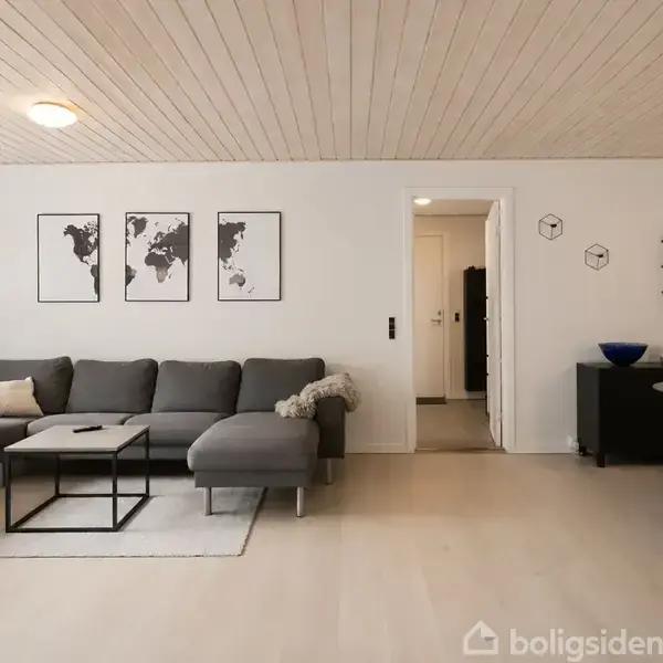 Grå sofa i stuen står foran billeder af verdenskort, ved siden af et lille bord og et spiseområde. Lyse trægulve og loft, med minimalistisk indretning.