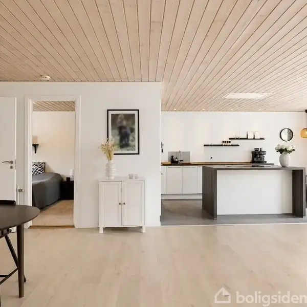 En åben stue designet i lyse nuancer; spisebord står i forgrunden, mens et moderne køkken med ø-bar i baggrunden. Store vinduer slipper dagslys ind.
