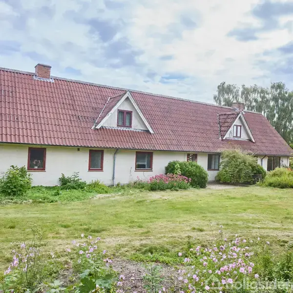 En hvid bondegård med rødt tegltag står stille omgivet af en velplejet have med buske og blomsterbede. Himlen er let overskyet.