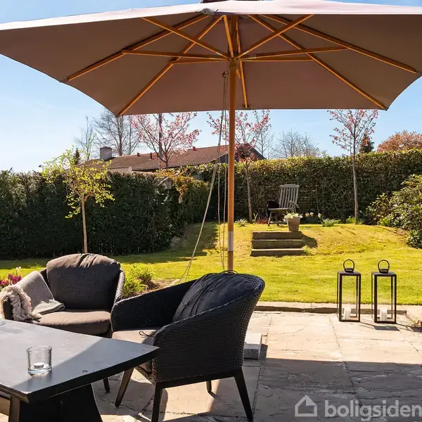 En terrasse med stole og bord under en stor parasol. I baggrunden er der en velplejet have med græsplæne, hækkesektioner, og blomstrende træer under en klar blå himmel.
