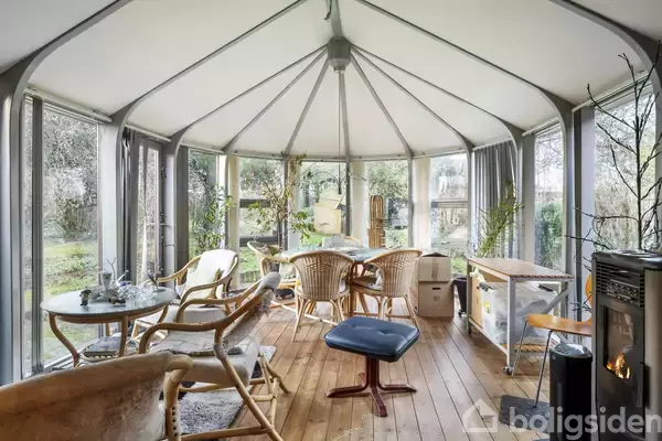 Vinterhave med rattanmøbler står omkring et rundt bord på et trægulv. Pejsen i hjørnet skaber en hyggelig atmosfære, omgivet af store vinduer med udsigt til haven.