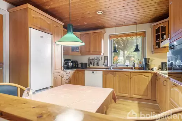 Køkken med træskabe og bordplade. Køleskab til venstre. Bord med lys dug står centralt. Grønne lamper hænger over bordet. Vindue giver udsigt til have med træer.