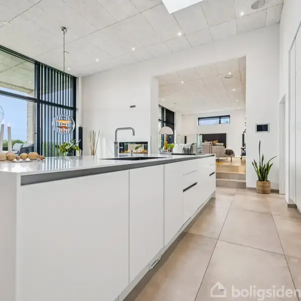 Køkkenø med hvidt kabinetarbejde står i et moderne køkken, omgivet af store vinduer. Overfladen har en vask, dekorationer og belysning. Stuen ses i baggrunden gennem en åben passage.