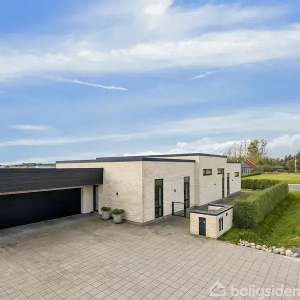 Moderne enfamilieshus med fladt tag står stille på en flisebelagt indkørsel med grønne buske og åben vej i baggrunden under en blå himmel.