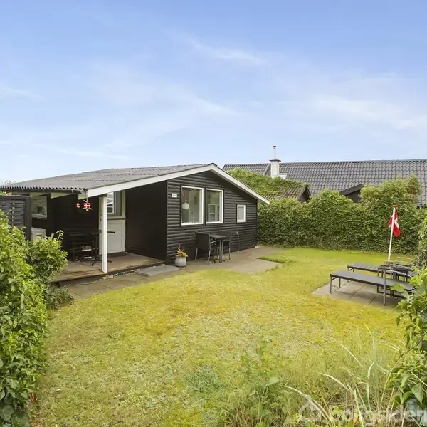 Et sort sommerhus med et lille overhæng over terrassen står i en grøn have. En grussti fører til indgangen. Et dannebrogsflag pryder en picnic-bænk i græsset.