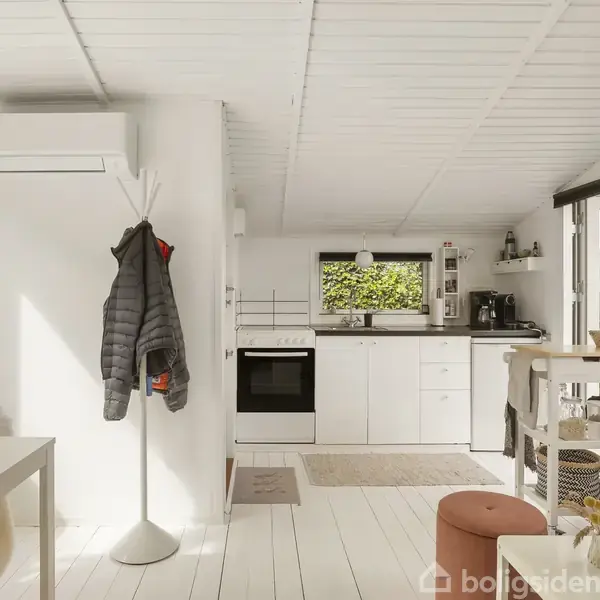 Køkken-alrum med hvidt interiør, hvor en jakke hænger på en stumtjener. Moderne køkken i baggrunden med hvide skabe og komfur. Lyse gulve og et vindue mod en grøn udsigt.