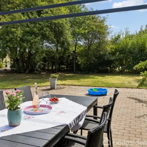 Et sort havebord med stole står på en terrasse, dækket med dug, blomster og en flaske. I baggrunden ses en grøn have med træer og en blå børnepool.