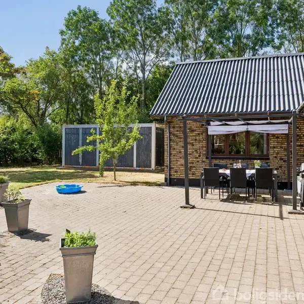 En brolagt terrasse med havemøbler og grill står foran en gulstensbygning. Omkring er der grønne træer og buske, med en lille blå plastikpool og plantekrukker spredt i haven.