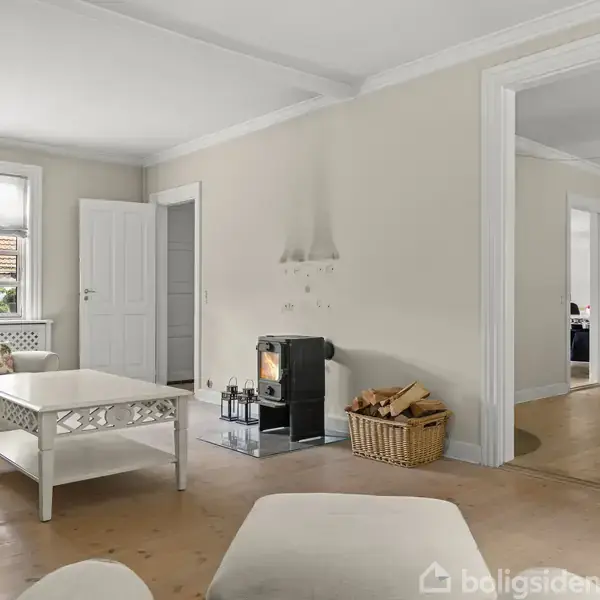 Brændeovn opvarmer stue med lyse vægge og lyse trægulve. Omkring står en kurv med brænde, sofaer, sofabord og en lampe. Store vinduer og en åben dør til næste rum.