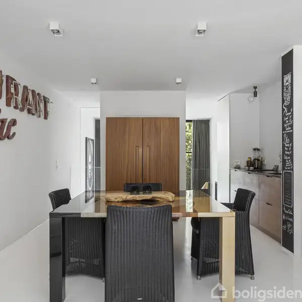 Et spisebord med sorte stole står i et minimalistisk rum med hvidt loft og vægge, træskab, og en tavlevæg med opskrifter og lister. På væggen ses bogstaverne "RANT".