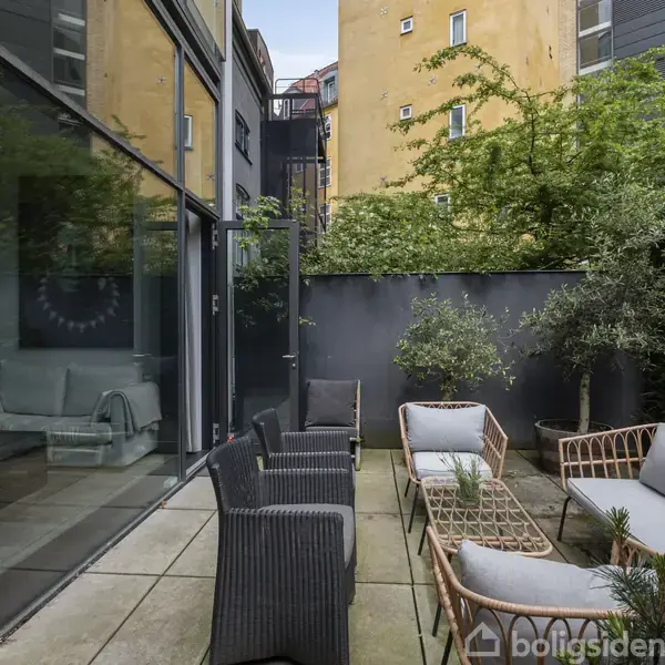 Udendørs loungeområde med flettede møbler og puder placeret på fliser. Høje glasvinduer reflekterer møbler, mens grønne planter og træer omkranser området i en bymæssig gårdhave.