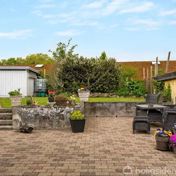 Terrasse med flisebelægning og potteplanter, nogle blomstrende. Haven har en hvid bygning og grønne planter i baggrunden. Et bord med to stole står ved en lysegul mur.