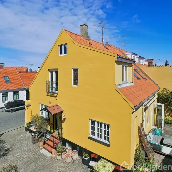 Gult hus står i sollys, med orange tegltag og balkoner, omgivet af brostensbelagt gård fyldt med planter i potter. Nabohuse med røde tage er synlige under blå himmel.