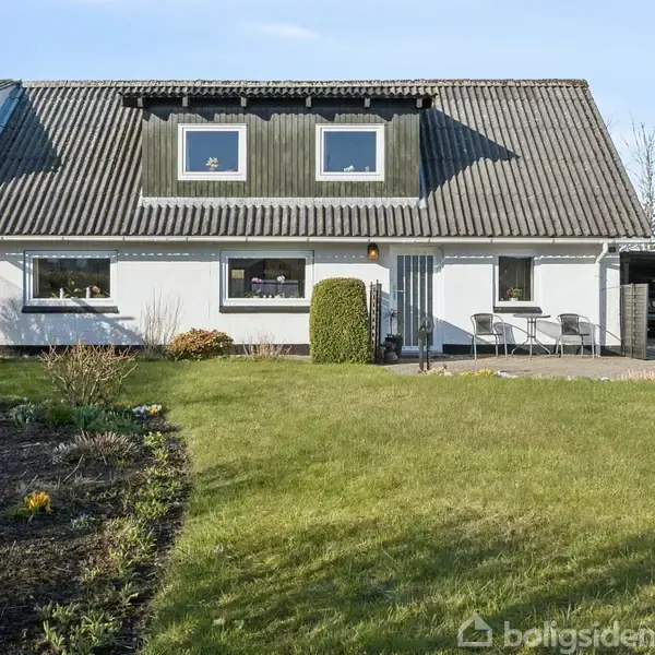 Hus med mørkt tag og tre kvistvinduer, står roligt foran en velholdt have. Til venstre er der blomsterbede, og til højre en indkørsel med carport.